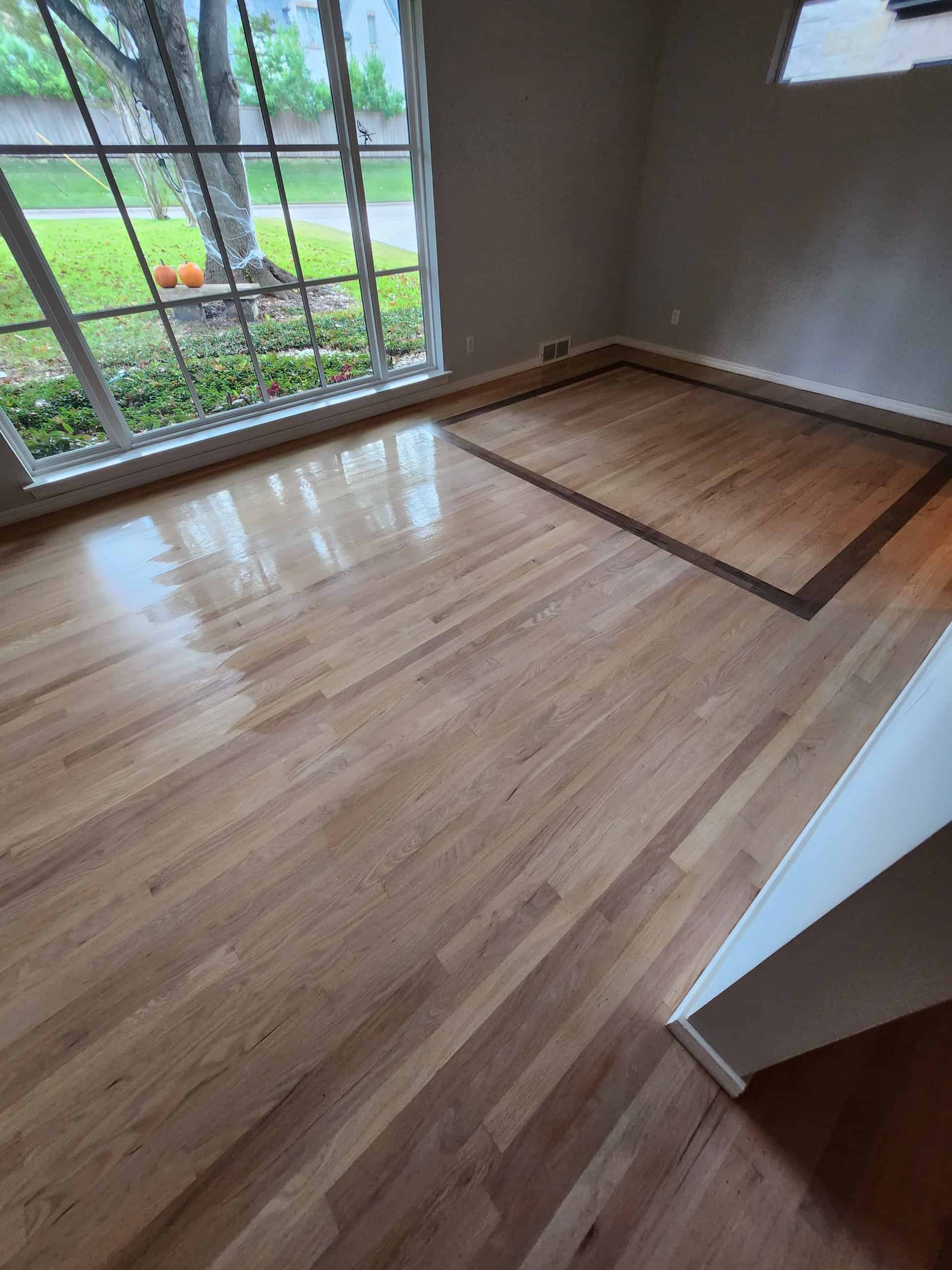 A bright room with polished hardwood floors featuring a darker wood border section. Large windows let in natural light, revealing a view of a green lawn with pumpkins outside. Walls are light-colored.